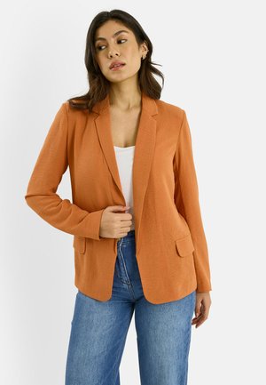 Mujer con blazer texturizado naranja sobre una camiseta blanca y jeans azules, posando con una mano cerca de la abertura del blazer.