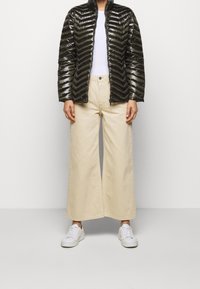 Sort pufferjakke med chevronmønster, hvid t-shirt, beige brede bukser og hvide sneakers på en neutral baggrund.