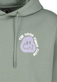 Groene hoodie van zacht materiaal met een paarse blobafbeelding en de tekst "THE GOOD VIBES CLUB" in het wit. Koord in de halslijn.