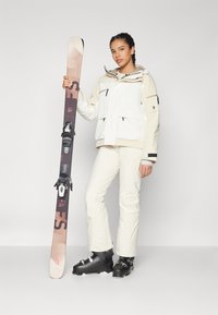 Skioutfit med en hvid isoleret jakke med sorte detaljer, matchende hvide bukser og sorte skistøvler. Holder en brun og pink ski.