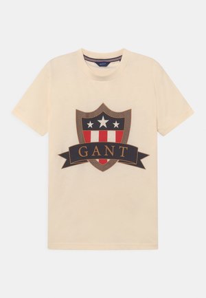 Beige T-shirt med korte ærmer med et skjoldemblem med stjerner og striber og et bånd med teksten "GANT" på tværs af brystet.