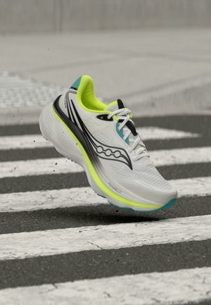 Chaussure de course blanche et vert néon avec des accents noirs et bleu sarcelle en l'air au-dessus d'un passage piéton sur une rue asphaltée.