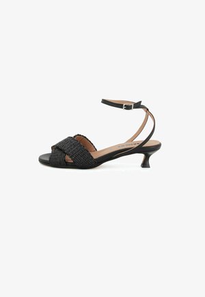 Sandalo nero con tacco basso, cinturini intrecciati anteriori, cinturino alla caviglia con fibbia e soletta imbottita beige su sfondo bianco.