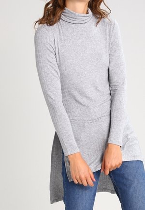 Kobieta w jasnoszarej prążkowanej tunice z golfem i długim rękawem oraz niebieskich dżinsach, pozująca z rękami przy udach.