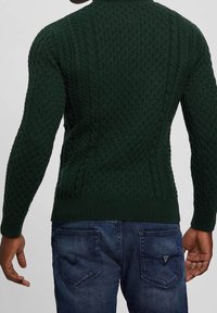 Dark green knitted sweater featuring a cable pattern, ribbed cuff and hem, paired with blue denim jeans.
