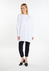 Losse witte trui met lange mouwen, ronde halslijn en een hoge-lage zoom, gecombineerd met zwarte cropped broek en zwarte instappers.