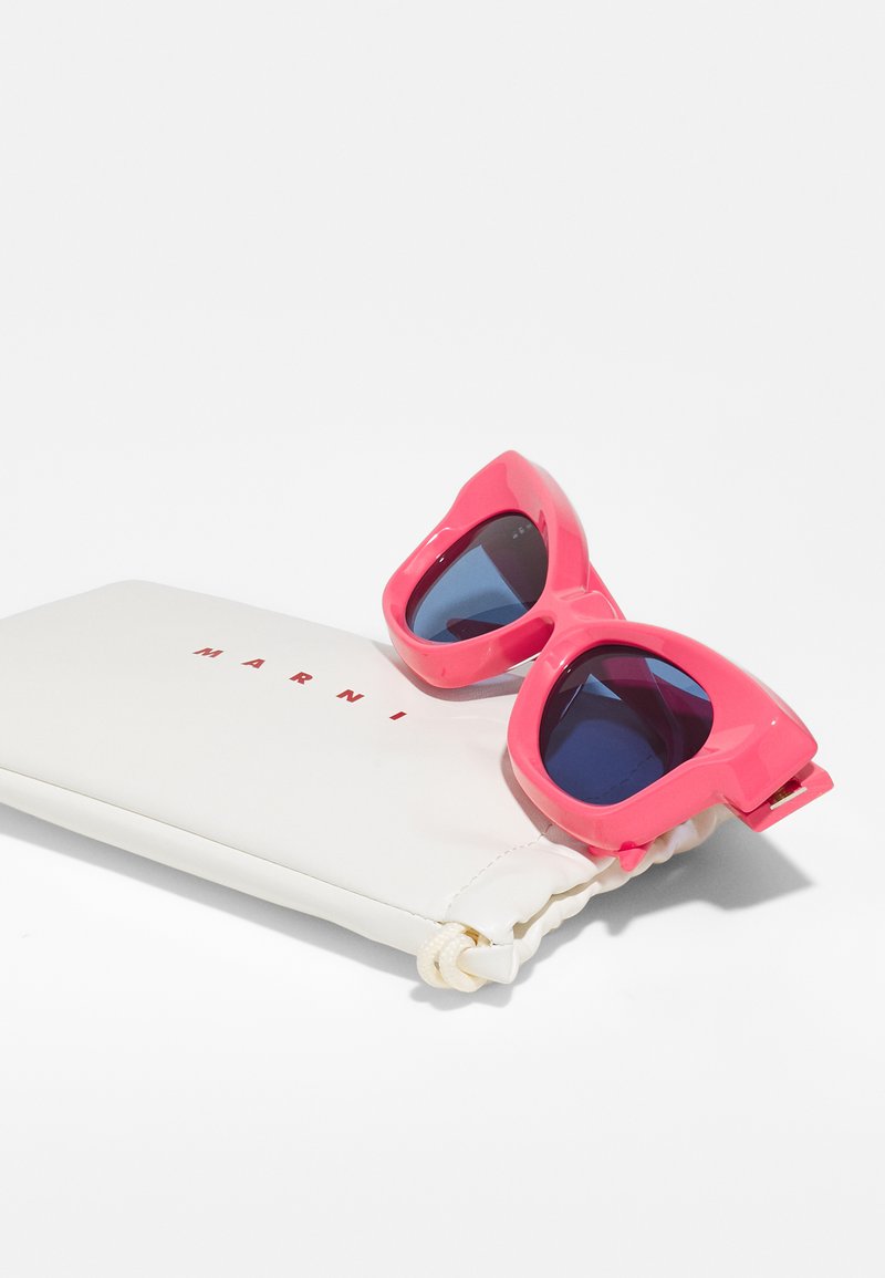 Gafas de sol de plástico rosa con lentes oscuros reposando sobre un bolso blanco, con el nombre de la marca "MARNI" en letras rojas.