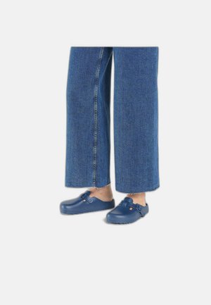 Zoccoli blu slip-on con cinturino regolabile, realizzati in materiale liscio. Abbinati a jeans denim a gamba larga con orlo sfrangiato.