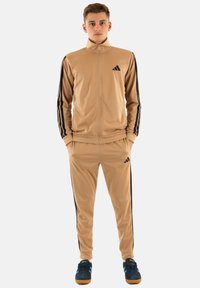 Giovane uomo in piedi con le mani nelle tasche, indossa una tuta beige con strisce nere e logo nero, sneakers blu navy con suole marroni.