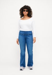 Jeans de mezclilla azul acampanados con una cintura alta, combinados con una blusa blanca de manga larga. La modelo lleva gafas de sol negras y zapatillas con estampado.