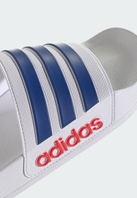 White slide sandal featuring three blue stripes, textured strap, and red "adidas" logo. Smooth material with a contoured footbed.