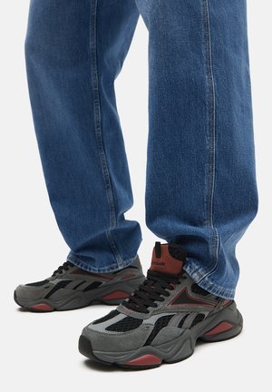 Reebok superge v sivi barvi z črno mrežasto tkanino in rdečimi poudarki, ki imajo zaobljen toe, debel podplat in kontrastne teksture. Jeans so modri denim.