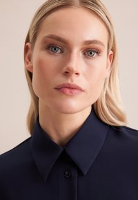 Camicia blu navy con colletto classico, chiusura a bottoni e texture liscia. Presenta una vestibilità su misura e una leggera lucentezza.