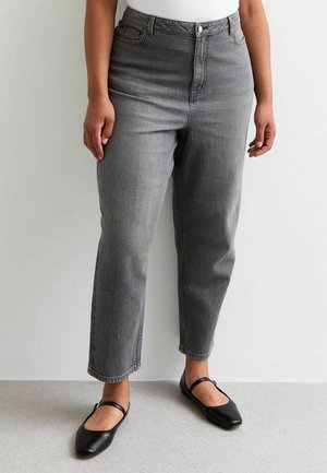 Osoba ubrana w szare dżinsy z wysokim stanem i czarne baleriny typu Mary Jane, stojąca na tle gładkiej, jasnej ściany.