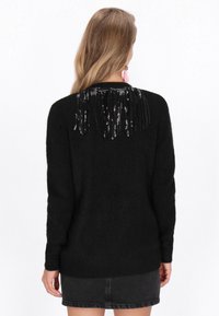 Fekete kötött pulóver, amely texturált hátrésszel, fekete rojttal és flitterekkel díszített. Hosszú ujjú, bordázott mandzsettával rendelkezik.