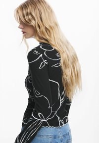 Black ribbed long-sleeve top with white abstract stitching, featuring a scalloped hem and fitted design, paired with light denim jeans.