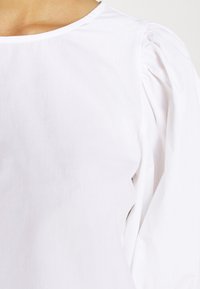 Bílá blůza s kulatým výstřihem a nabíranými rukávy. Vyrobena z lehkého materiálu s hladkou texturou. Bez viditelných vzorů nebo akcentů.