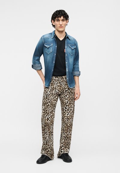 Νεαρός άνδρας που στέκεται, φοράει τζιν πουκάμισο με τα μανίκια γυρισμένα πάνω από μαύρο μπλουζάκι, παντελόνι με λεοπάρ μοτίβο και μαύρα παπούτσια, μπροστά σε απλό ανοιχτόχρωμο φόντο.