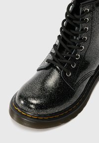 Botín de tobillo negro brillantoso con un acabado lustroso, que presenta cordones negros, ojetes plateados y una suela gruesa negra con un acento amarillo alrededor del borde.