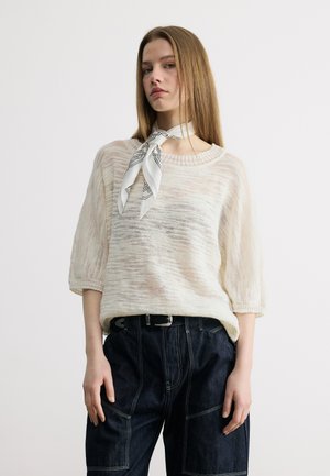 Mujer que lleva un suéter de punto blanco transparente con mangas hasta el codo, una bufanda blanca con estampado en el cuello y jeans oscuros de talle alto con costuras en contraste.