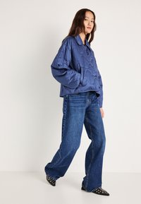 Κουβερτίνα ναυτικού χρώματος μπουφάν με στρογγυλή γιακά και κλείσιμο με κουμπί, συνδυασμένο με μπλε ψηλόμεσα τζιν και μαύρες παπούτσια με μυτερές μύτες και τρουκς.