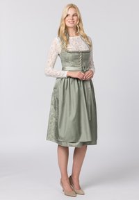 Grünes Dirndl mit tailliertem Oberteil, langen Spitzenärmel und filigranen Blumenmustern. Verfügt über Knöpfe und eine gebundene Taille, kombiniert mit beigen Absätzen.