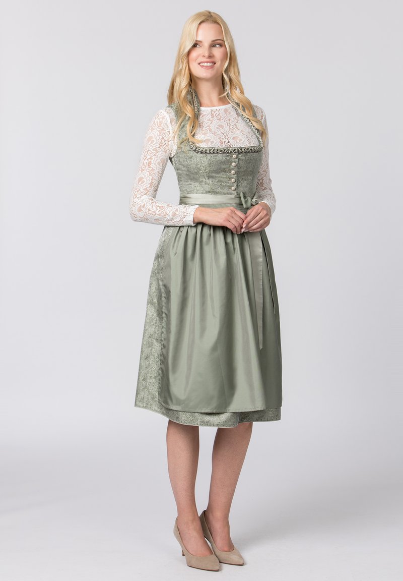 Grünes Dirndl mit tailliertem Oberteil, langen Spitzenärmel und filigranen Blumenmustern. Verfügt über Knöpfe und eine gebundene Taille, kombiniert mit beigen Absätzen.