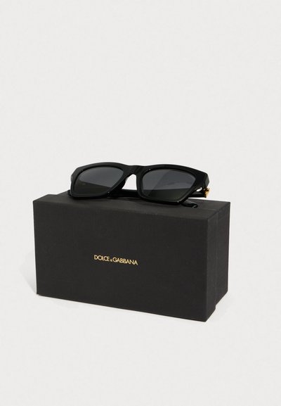 Čierne slnečné okuliare s širokým, obdĺžnikovým rámom ležiace na čiernej krabici s zlatým brandingom "Dolce & Gabbana". Skle sú tmavé.