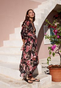 Blommig kimono i svart med rosa och röda blommor, flödande tyg, midjebälte, öppen framstil, kombinerad med svarta flip-flops, mot en ljus bakgrund.