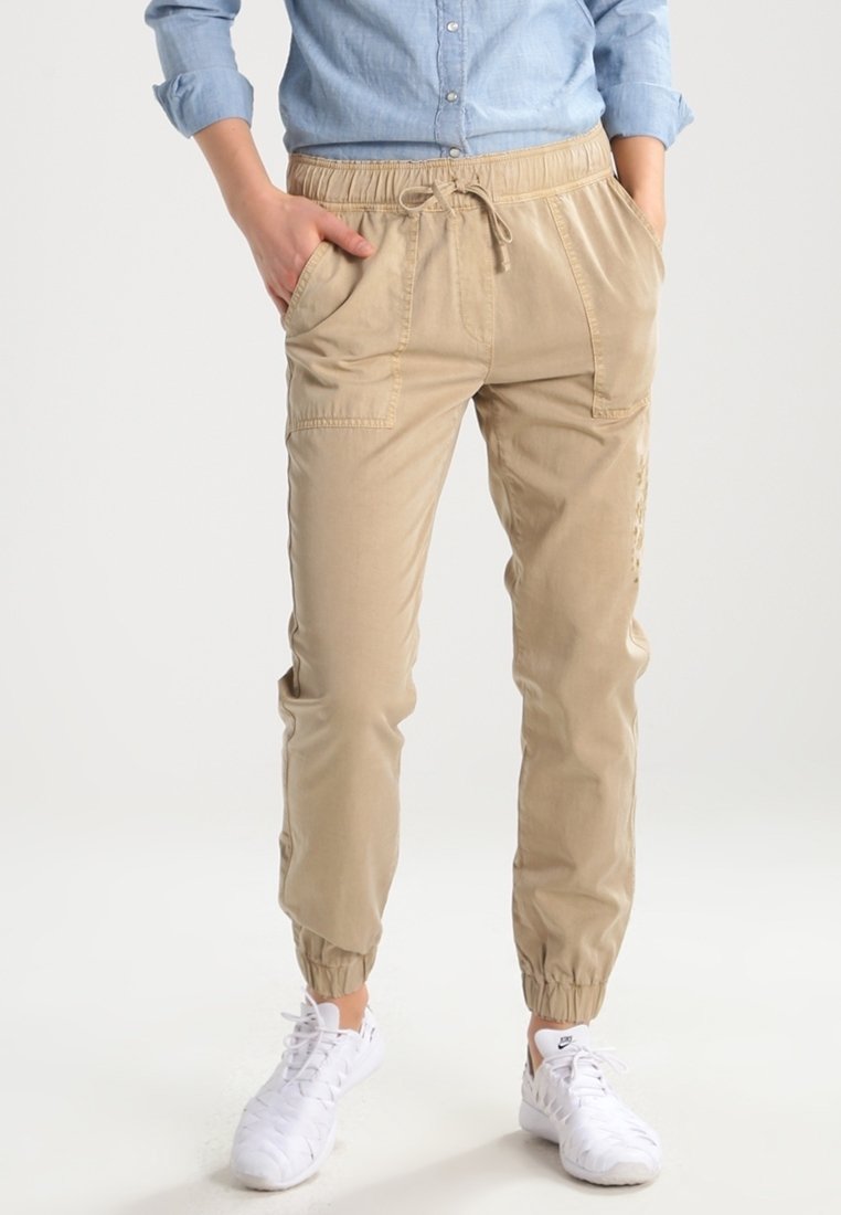 Personne portant un pantalon de jogging beige à cordon avec des poignets élastiques, une chemise bleu clair boutonnée aux manches retroussées, et des chaussures de sport blanches.