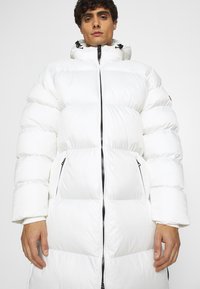 Cappotto imbottito bianco con colletto alto, caratterizzato da una chiusura con zip anteriore, ampie sezioni trapuntate e due tasche laterali conzip.
