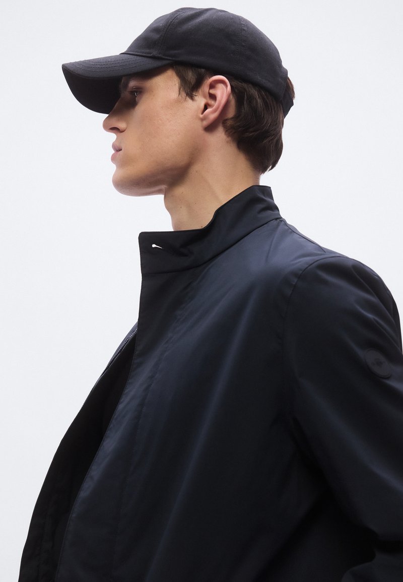 Black jacket with high collar and zip closure, paired with a black cap. Smooth texture and streamlined design, featuring subtle branding accents.