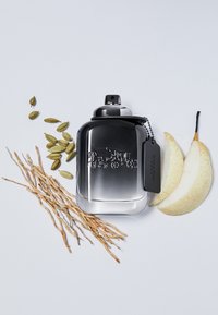 Flacon de parfum noir en forme arrondie avec un design gravé, entouré de gousses de cardamome, de racines de plantes et de tranches de poire.