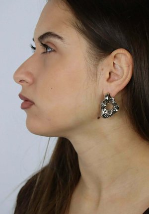 Orecchini metallici con una forma ondulata e organica con una finitura testurizzata, indossati all'orecchio di una modella. Il colore appare argento con un aspetto lucido.