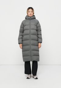 Dikke grijze lange jas met een capuchon, voorzien van een ritssluiting aan de voorkant, zijzakken en een gewatteerd ontwerp. Gedragen over zwarte broek en schoenen.