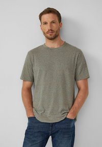 Gestreiftes, kurzärmliges T-Shirt in gedämpftem Grün und marineblau, aus Baumwolle gefertigt. Runder Halsausschnitt und lockere Passform, kombiniert mit dunkelblauen Jeans.