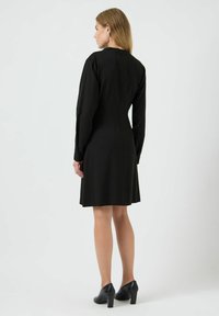 Černé šaty s dlouhými rukávy, přiléhavým tělem a rozšířenou sukni. Zapínání na zip na zadní straně. Nosené s černými lodičkami. Hladká textura látky.