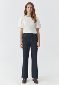 Weißes Oberteil mit schulterlangen Ärmeln, dunkelblaue High-Waisted Schlagjeans, braune Stiefeletten, glatte Texturen und lässiges Design.