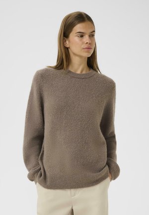 Brun fluffy sweater med rund halsudskæring, lange ærmer og ribbede manchetter, båret over lyse bukser mod en hvid baggrund.