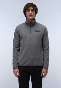 Sudadera de felpa gris con cuello con cierre de cuarto, que presenta un pequeño logo en el pecho y una textura suave. Lleva puesta con pantalones negros.
