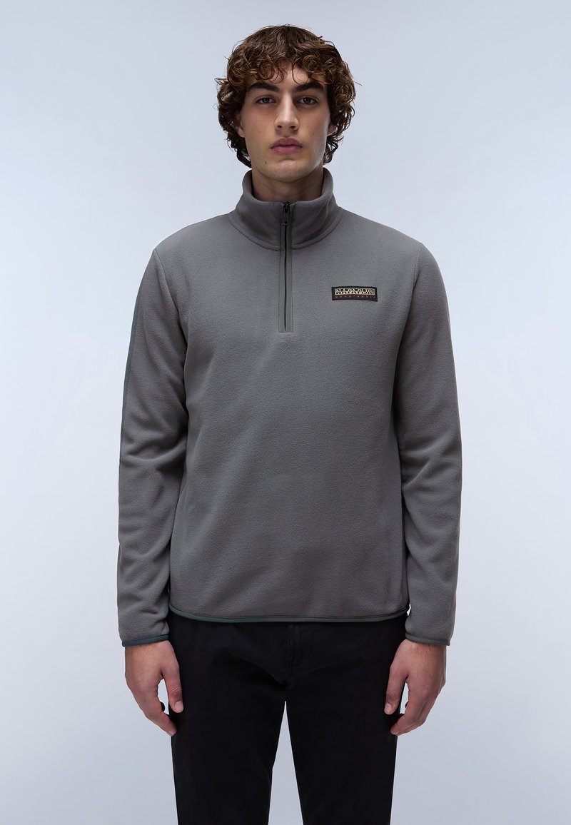 Sudadera de felpa gris con cuello con cierre de cuarto, que presenta un pequeño logo en el pecho y una textura suave. Lleva puesta con pantalones negros.