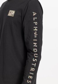 Camisa negra de manga larga hecha de algodón. Presenta un parche de camuflaje en el pecho y el texto "ALPHA INDUSTRIES" en impresión multicolor en la manga.