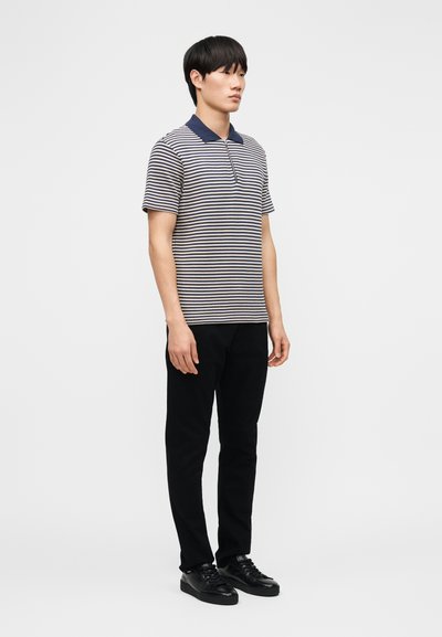 Άνδρας που στέκεται φορώντας ριγέ κοντομάνικο πόλο με ναυτικό γιακά, μαύρο παντελόνι και μαύρα αθλητικά παπούτσια, μπροστά σε ένα απλό φωτεινό φόντο.