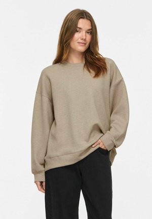 Jeune femme aux cheveux châtain clair portant un sweat-shirt beige oversize et un pantalon noir, debout avec une main dans sa poche.