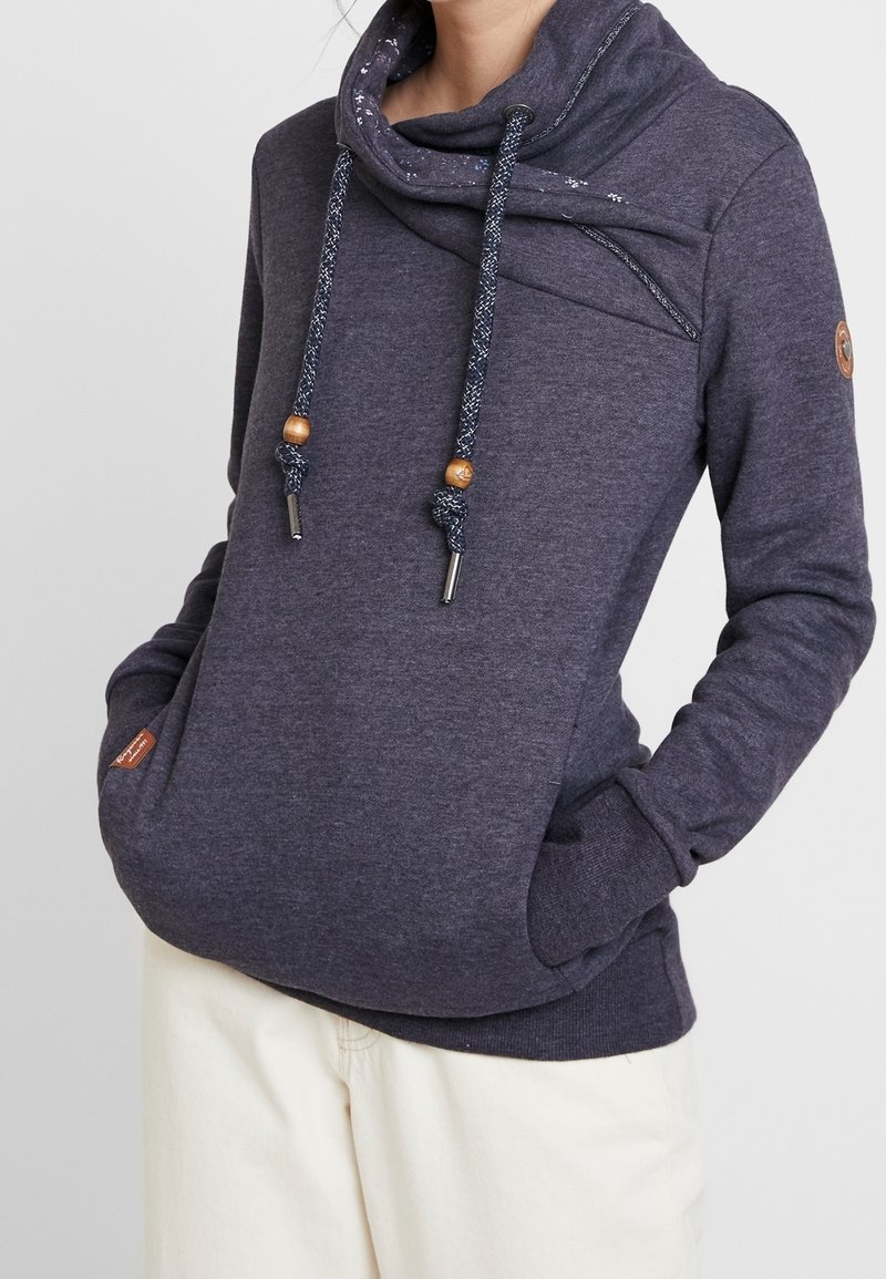 Personne portant un sweat à capuche bleu foncé avec un col haut et replié et des cordons décoratifs, mains dans les poches avant, associé à un pantalon clair.