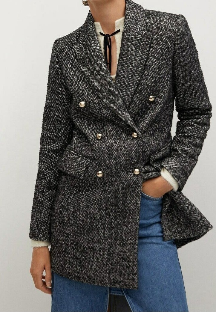 Personne portant un blazer à double boutonnage gris foncé texturé avec des boutons dorés, un haut blanc avec une cravate noire et un jean bleu.