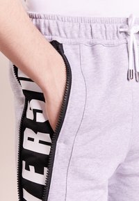 Ljusgrå sweatpants med en svart dragkedjeficka och kontrasterande textdesign. Tillverkade av mjukt tyg, med elastisk midja och dragsko.