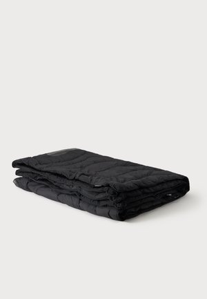 Couverture matelassée noire pliée soigneusement, présentée sur un fond gris clair uni.