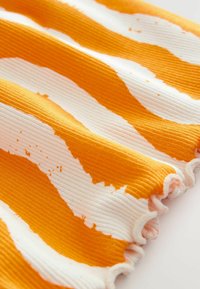 Orange-weißer gerippter Stoff mit wellenförmigen Mustern, leicht gekräuselter Textur und fransigen Kanten. Geeignet für Bekleidungsdesign.