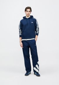 Μπλε σκούρο φούτερ με κουκούλα και λευκές ρίγες στα μανίκια, τσέπη καγκουρό, σετ με μπλε σκούρο παντελόνι με λευκές γραφικές λεπτομέρειες και μπλε αθλητικά παπούτσια.